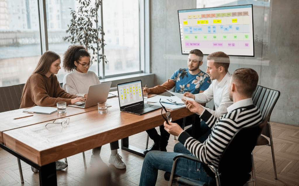 Five people gathered around a conference table in a modern office, collaborating with laptops, documents, and a shared screen displaying colorful charts.